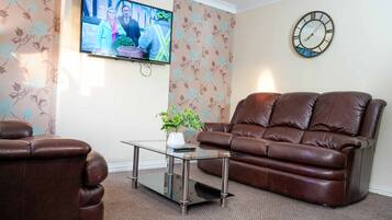A living room with a brown leather sofa, a glass coffee table, a wall-mounted TV, and a clock.