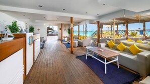Ruime, overdekte ruimte met een houten vloer, een bank met gele kussens en uitzicht op de zee door grote ramen.