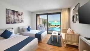 Hotelkamer met twee bedden, een kleine tafel, een rieten stoel en een televisie. De kamer heeft een groot raam met uitzicht op een zwembad en de zee.
