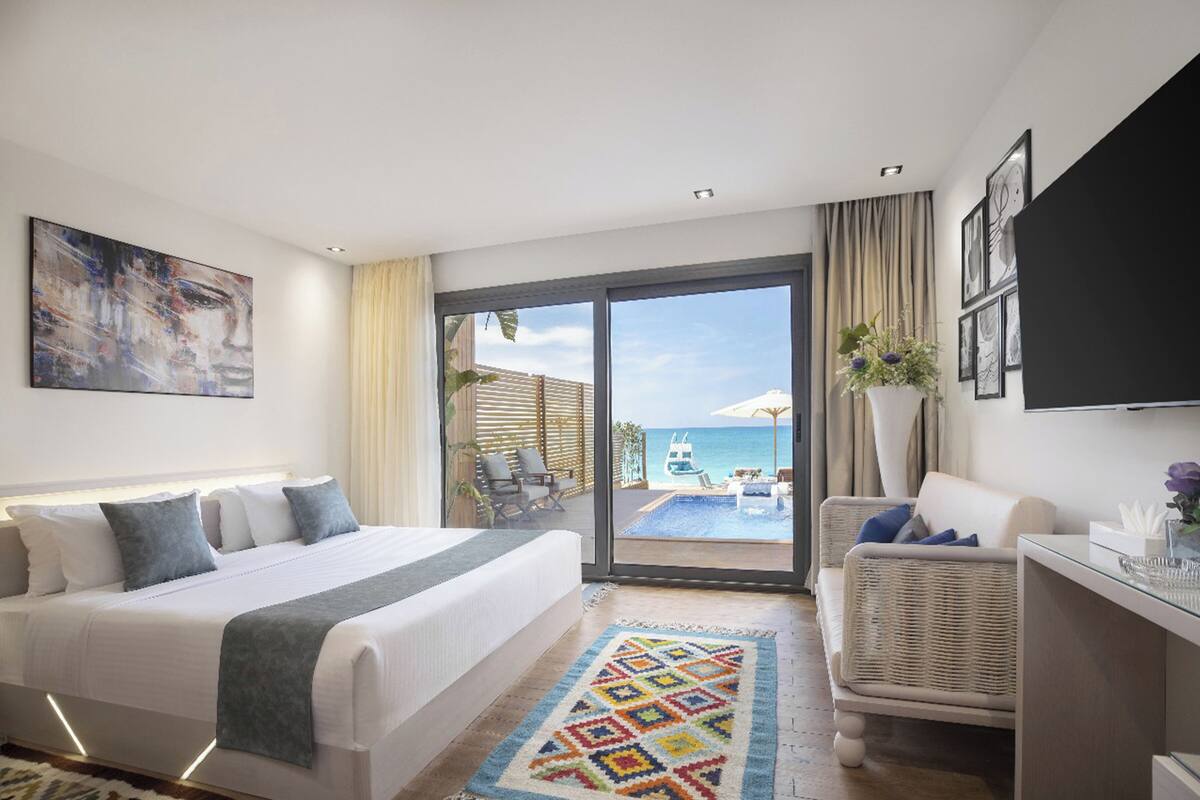 A hotel room with a bed, a sofa, a TV, a window with a view of the sea, and a balcony with outdoor furniture.