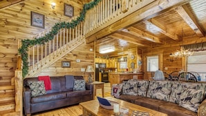 Interior de uma cabana de madeira com escada, sala de estar com sofá e mesa de centro, e área de cozinha.