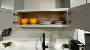 A kitchen with a white countertop, a black faucet, a toaster, and a shelf above the sink holding yellow bowls and stacked plates. - Up Unit Studio in West CLE (Cleveland)