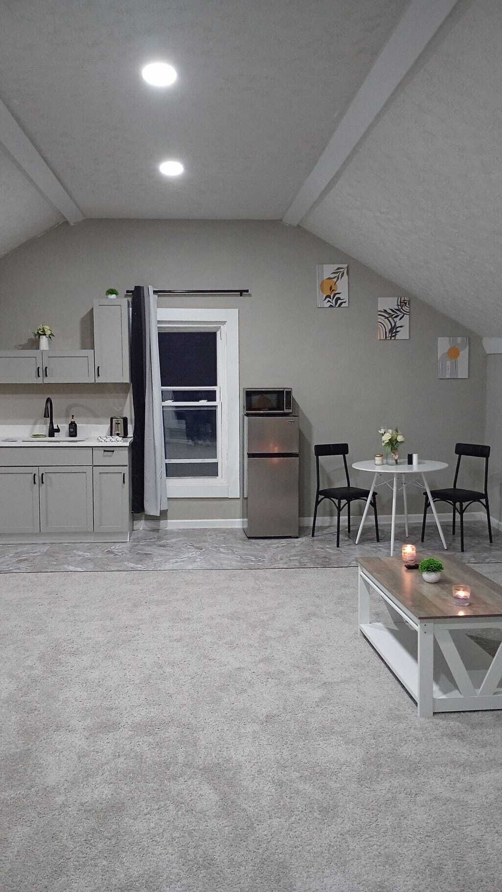 A compact kitchen with a refrigerator, microwave, and sink. There is a dining area with two chairs and a small table. The room has a sloped ceiling and a window.