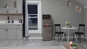 A compact kitchen with a refrigerator, microwave, and sink. There is a dining area with two chairs and a small table. The room has a sloped ceiling and a window.