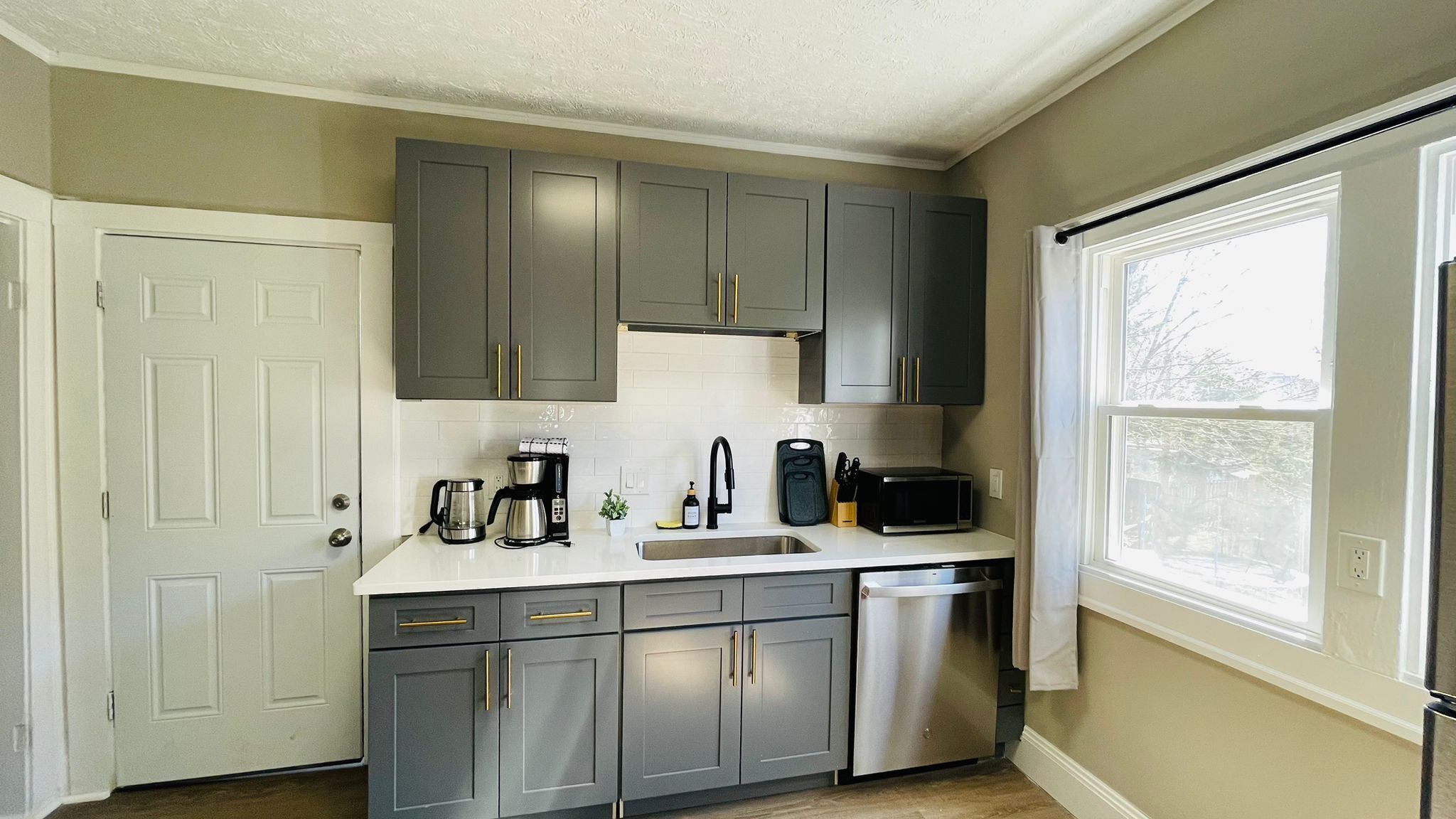 A modern kitchen with grey cabinets, a white countertop, a stainless steel dishwasher, and a window providing natural light.