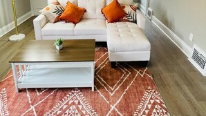 A living room with a white sofa, orange cushions, a coffee table, a floor lamp, and a painting on the wall. - The Stargazer Sunroom 2bedroom in West CLE (Cleveland)