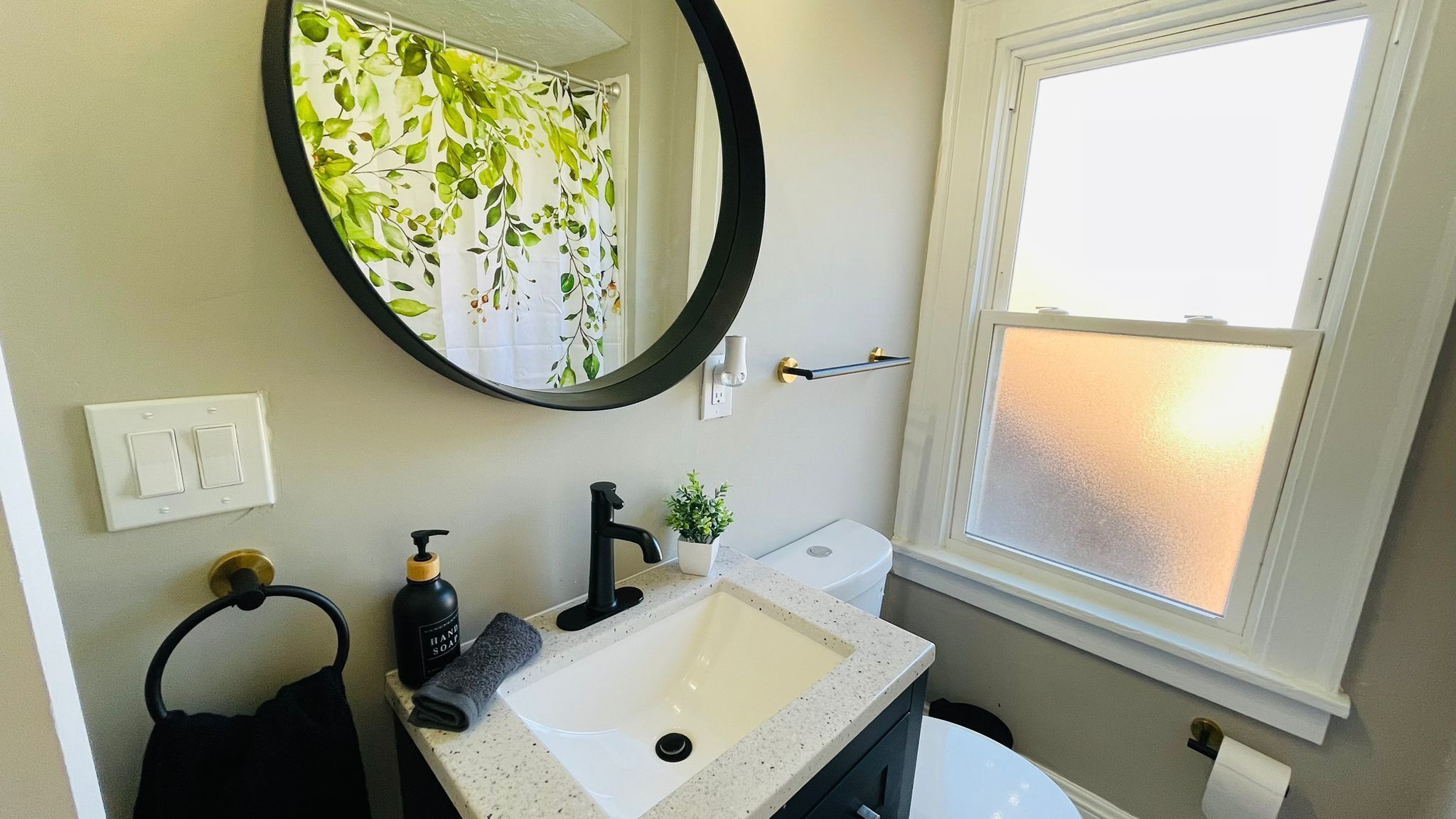A bathroom with a round mirror, a sink, a toilet, and a window with frosted glass.