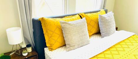 Una cama con ropa de cama estampada en amarillo y blanco, una mesita de noche con una lámpara, una planta pequeña y una botella de aceite de lavanda.