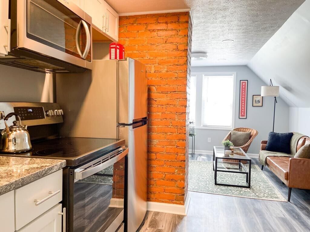 A kitchen with a microwave, stove, and refrigerator. There is a brick wall and a living area with a sofa and a coffee table.