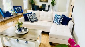 Een moderne woonkamer met een witte hoekbank, blauw-witte kussens, een glazen salontafel met decoratieve objecten en blauwe fauteuils.