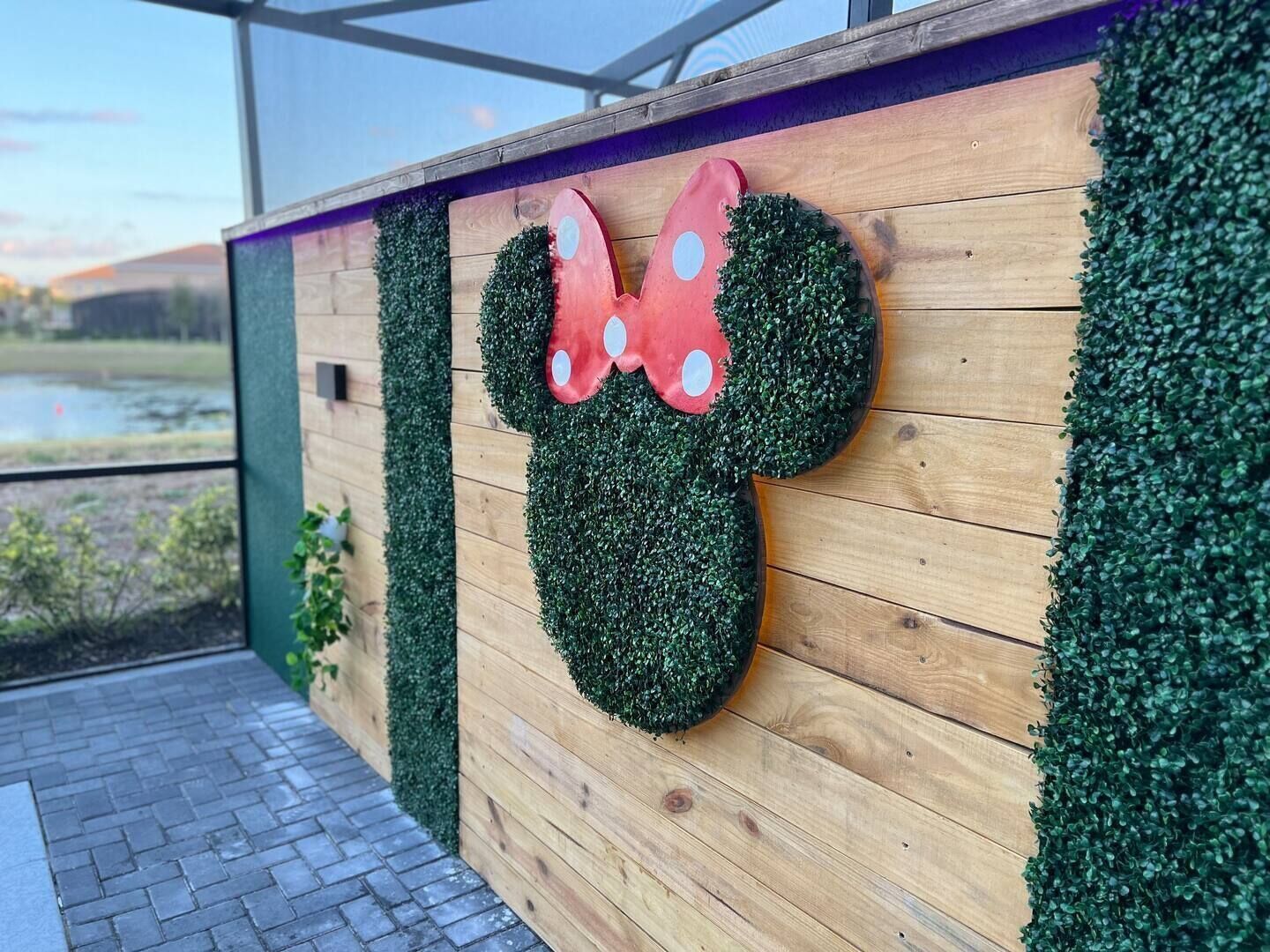 A wooden wall with a Mickey Mouse head made of green foliage and a red bow.