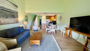 A living room with a sofa, coffee table, and TV. There is a dining area with chairs and a table. A kitchen is visible in the background. - Gold-rated 2 bed/2 bath condo with view of the lagoon, just steps to the pool. (Saint Augustine)