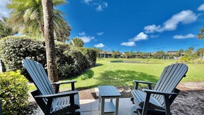 An outdoor seating area with Adirondack chairs and a small table, overlooking a well-maintained lawn and a distant building complex. - Awesome condo, steps to the heated indoor/outdoor pool, short walk to the beach! (Saint Augustine)