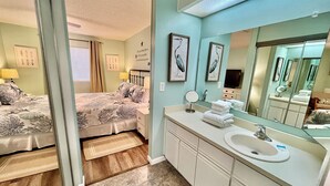 A bedroom with a bed, bedside table, lamp, and a bathroom with a sink, mirror, and towel stack. - Great property perfect for a couples getaway to the beach, steps to pool & sand! (Saint Augustine)
