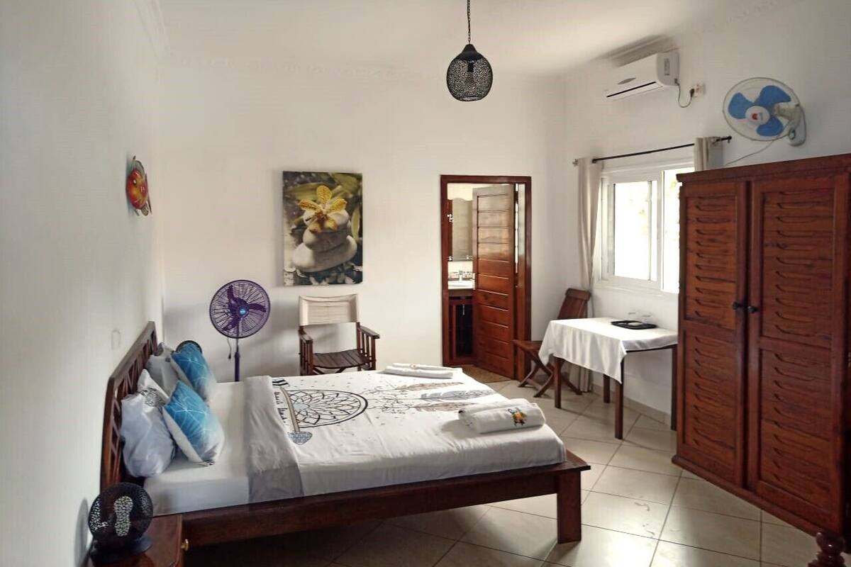A bedroom with a bed, a chair, a table, a wardrobe, and a fan.