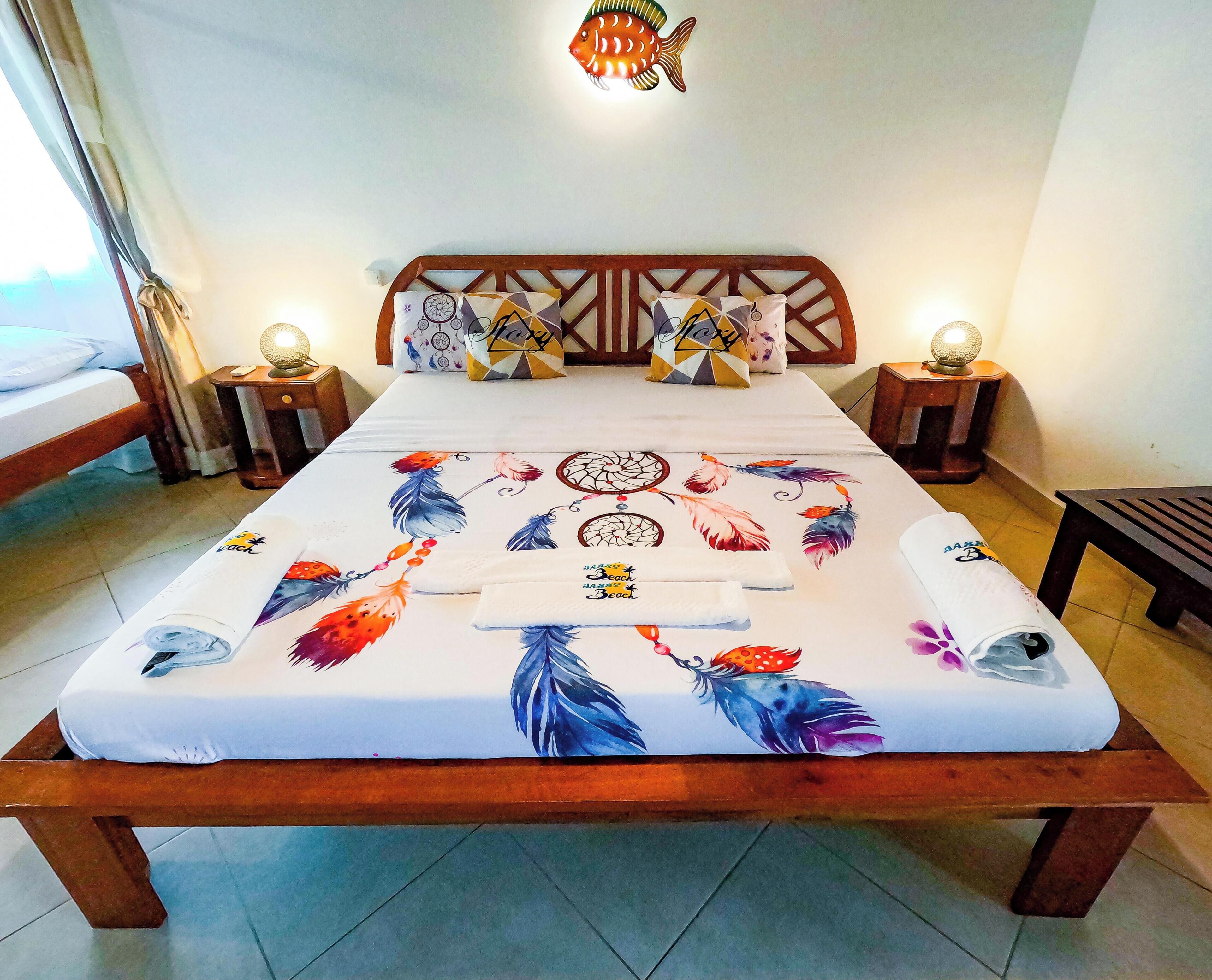 A neatly made bed with a colorful feather print bedspread, two bedside lamps, and a wooden headboard.