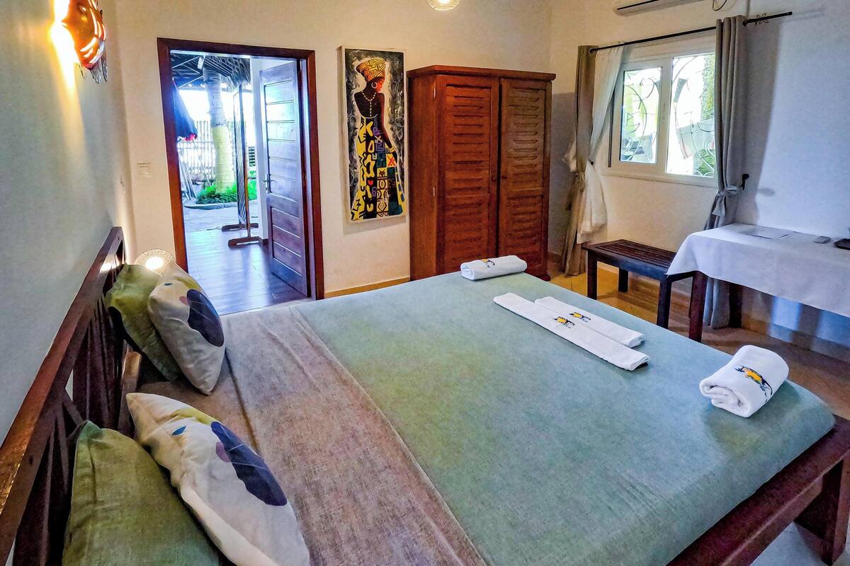 A hotel room with a bed, a ceiling fan, a painting, a wooden wardrobe, a table with a tablecloth, and a window with curtains.