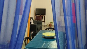 Une pièce avec une table à manger recouverte d’une nappe bleue, un ventilateur de plafond et un téléviseur posé sur un meuble.