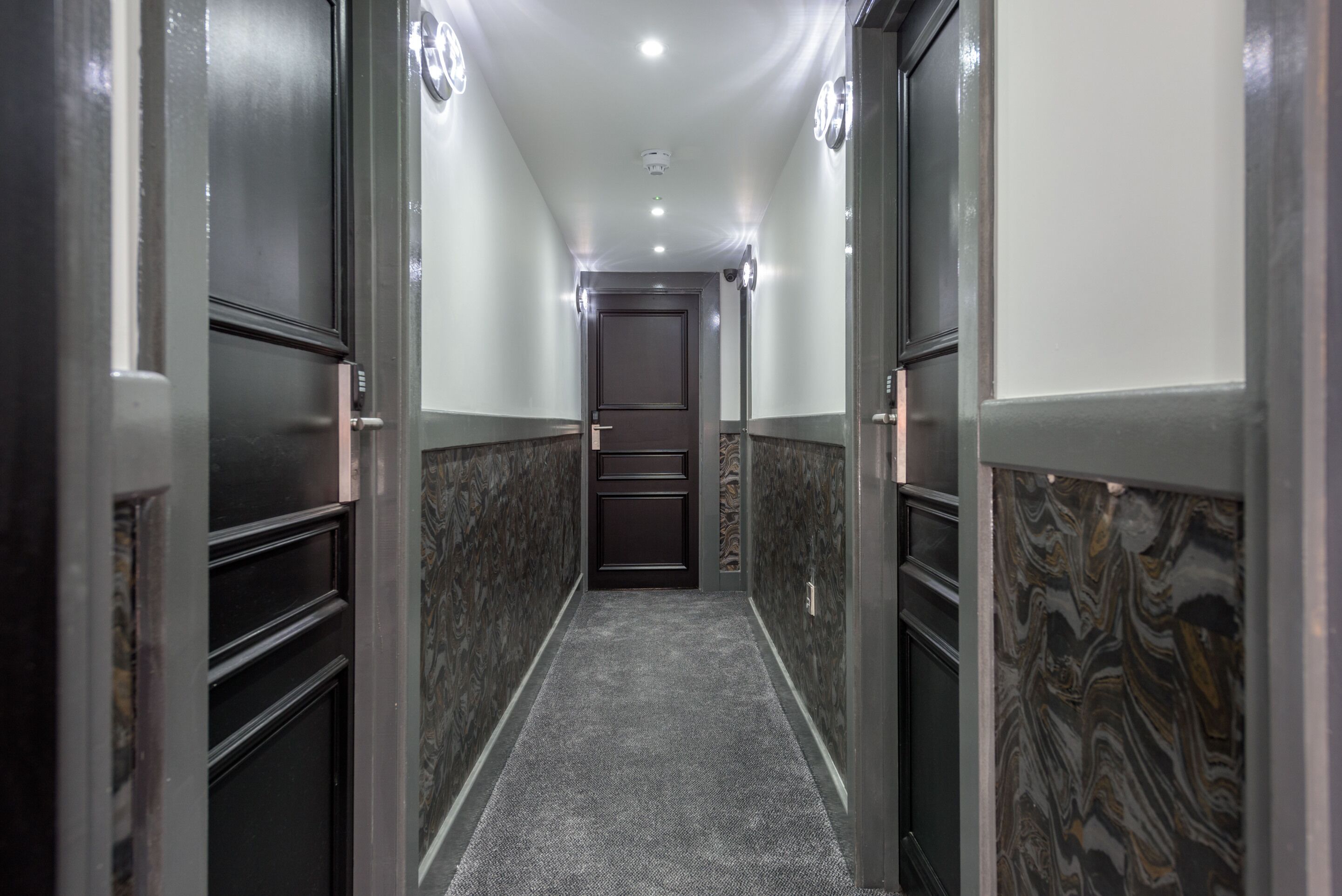A narrow hallway with a carpeted floor, dark wooden doors, and patterned wall tiles.