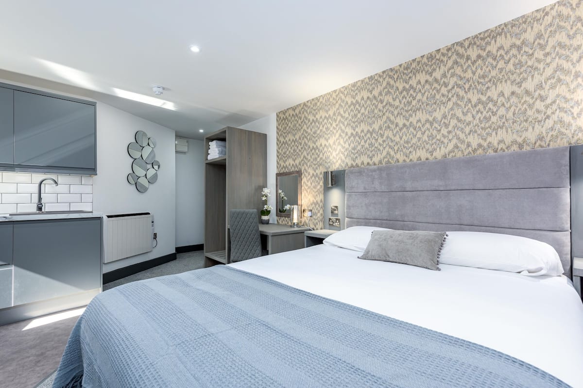 A modern hotel room with a large bed, a built-in desk, a vanity with a mirror, and a patterned wallpaper.