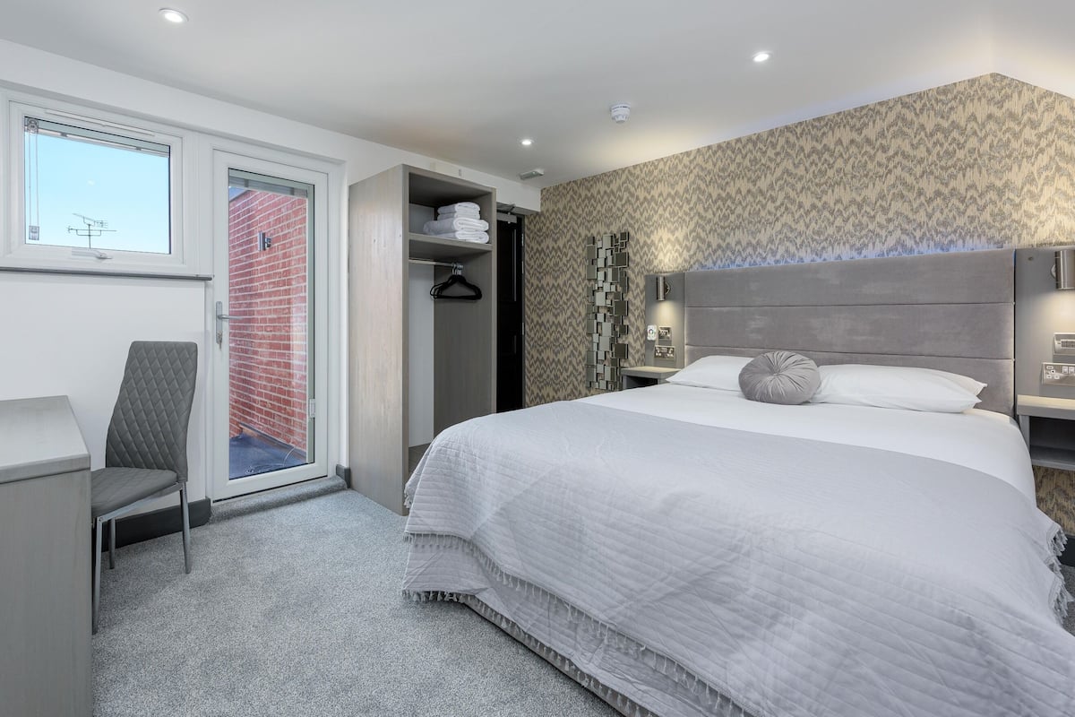 A modern hotel room with a large bed, a desk, a chair, a wardrobe, and a view of a brick building through a sliding door.
