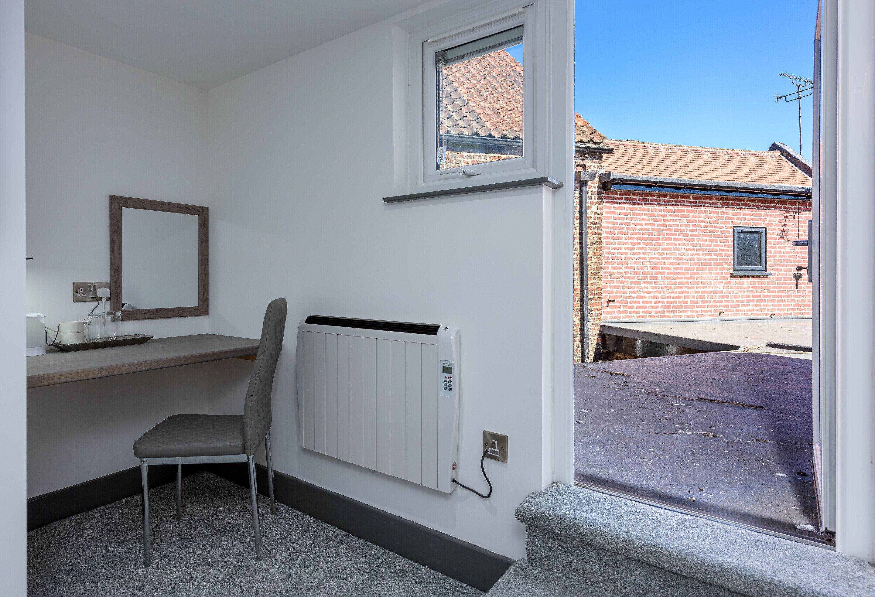 Una habitación con un radiador, una silla, una mesita, una ventana y vistas a un edificio de ladrillo.