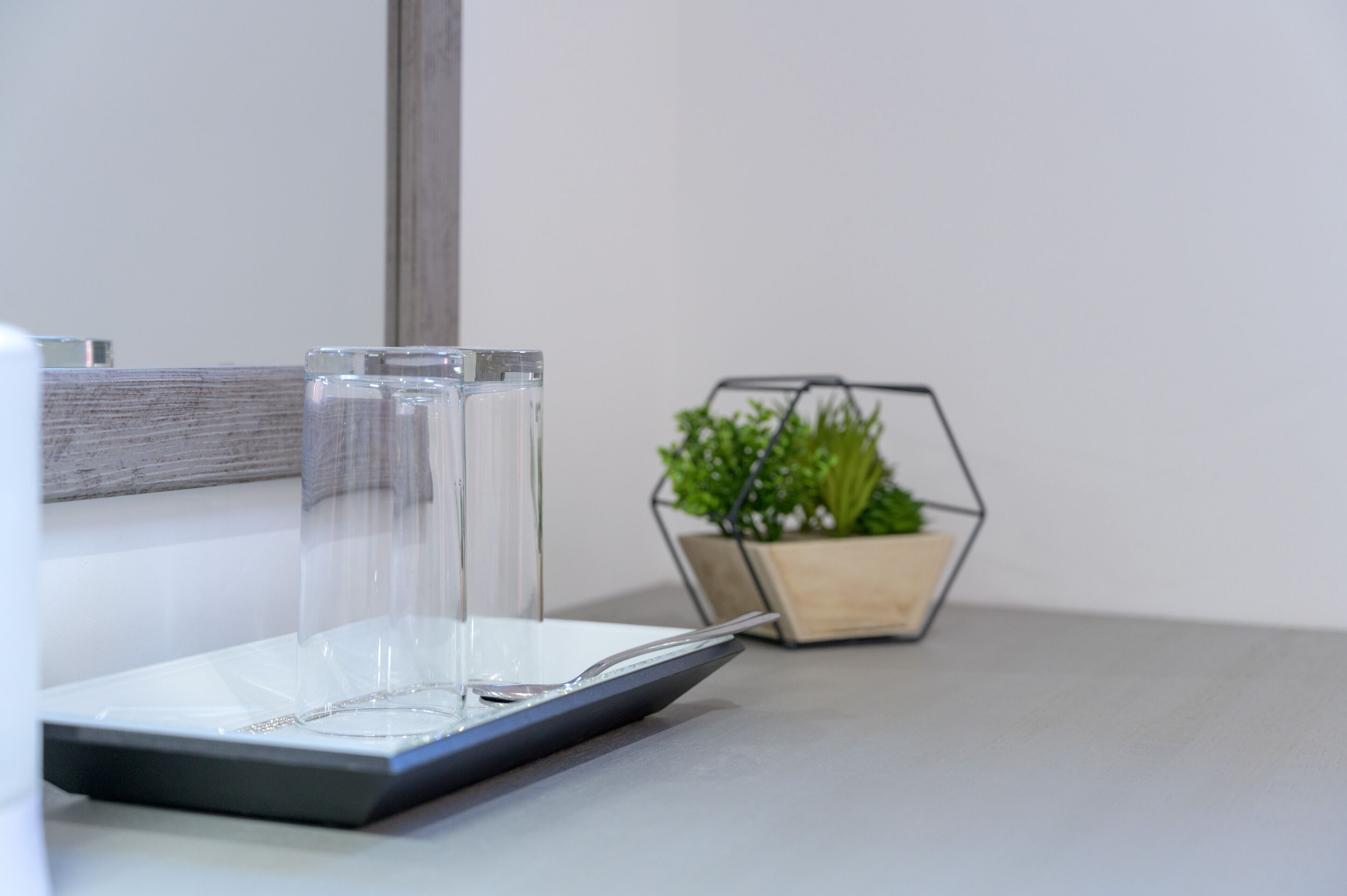 Um copo de vidro transparente sobre uma bandeja, uma colher e um vaso hexagonal com plantas verdes.