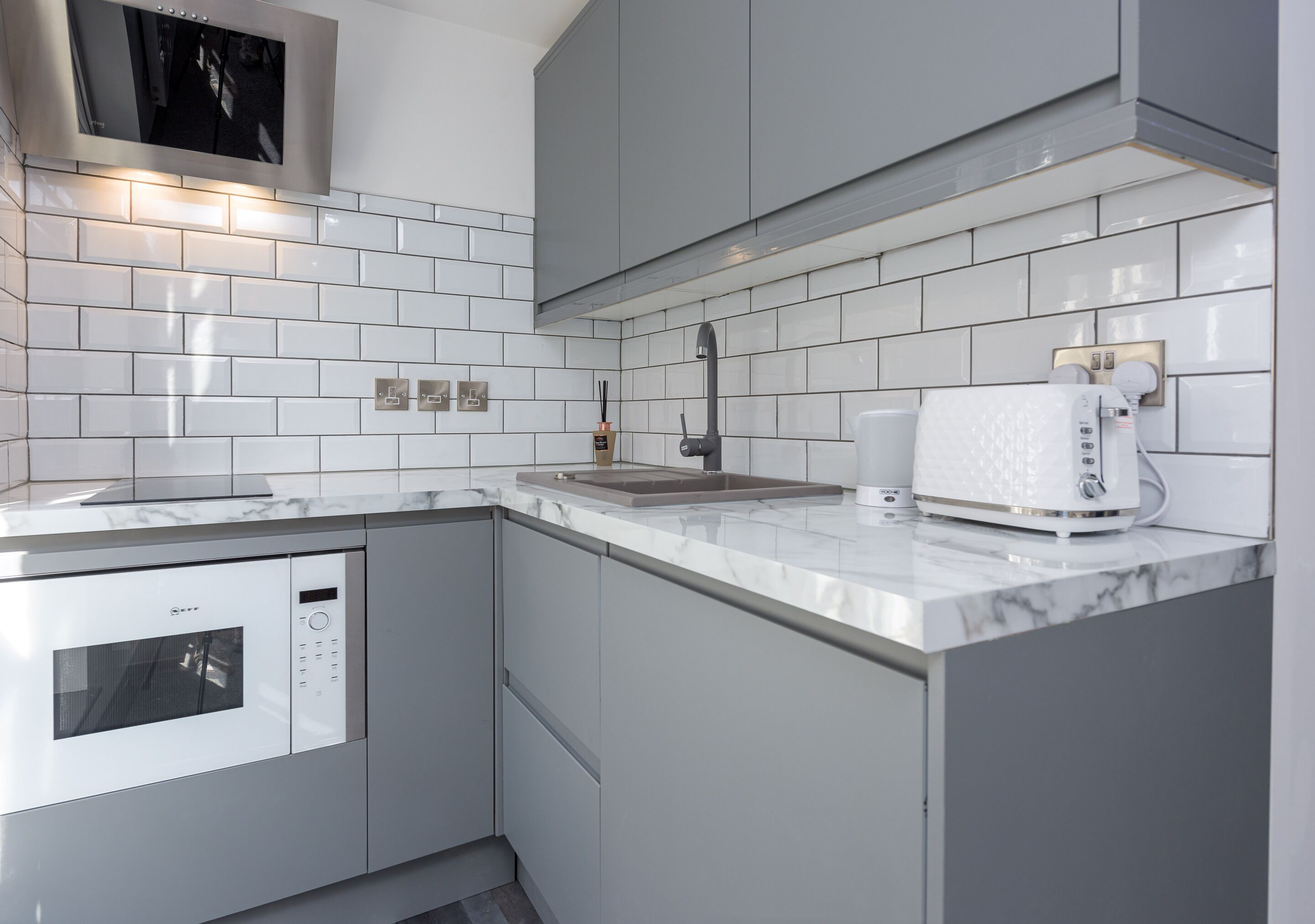 A modern kitchen with white subway tiles, a built-in microwave, a coffee maker, and grey cabinets.