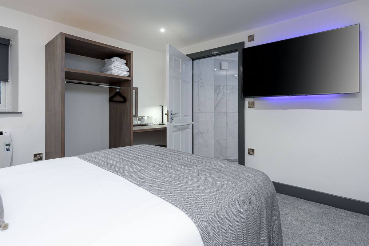 A modern hotel room with a bed, a wardrobe, a bathroom with a shower, and a mounted TV.