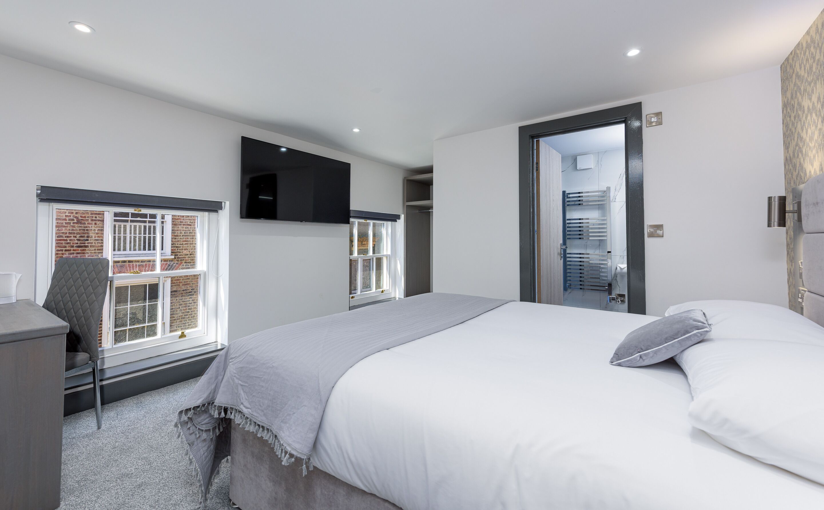 A modern bedroom with a bed, a TV mounted on the wall, a window with a view of a brick building, and a door leading to a bathroom.