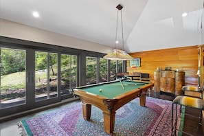 A pool table in a room with large windows, a hanging light fixture, and a wooden cabinet. - The Estate - Luxury with Pool Table & Game Room (South Pittsburg)