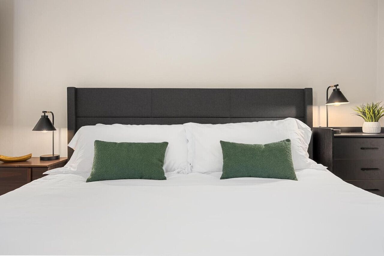 Un lit bien fait, avec du linge de lit blanc et deux coussins verts, flanqué de deux tables de chevet sur lesquelles sont posées des lampes, et sur l’une d’elles, une plante.