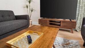 Una sala moderna con un sofá gris, una mesa de centro de madera con una bandeja que sostiene un control remoto, un televisor de pantalla plana sobre un mueble de madera, y una planta en maceta.