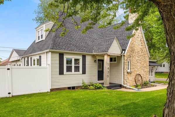 A two-story house with a stone chimney, a white picket fence, and a well-maintained lawn. - Suburban Sanctuary: 3BD Perfect for Long Stays (Pekin)