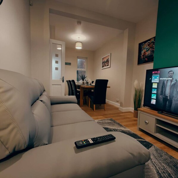 A living room with a grey sofa, a TV on a stand, a remote control on the sofa, a dining area in the background, and a green wall.