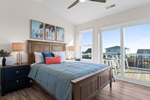 A bedroom with a bed, nightstands, a ceiling fan, artwork, and large windows with a view. - Enjoy stunning ocean views at Chipps Ahoy, a spacious 4-bedroom retreat in Oak Island. (Oak Island)
