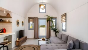 A modern living room with a grey sofa, a TV, and a kitchen area with a white countertop and a red kettle. - ACL'L Luxury Suites (Santorini)