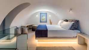 A modern bedroom with a curved ceiling, a bed with a blue accent, bedside tables, and a glass staircase. - ACL'L Luxury Suites (Santorini)