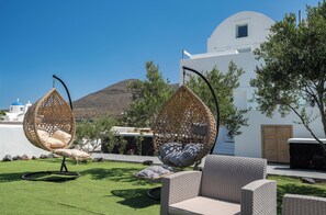 A patio with wicker chairs and a wicker swing chair, a white building, and a mountain in the background.
