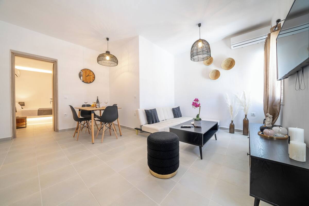 A modern living room with a sofa, dining table, and ottoman. There are hanging lights and a TV mounted on the wall.