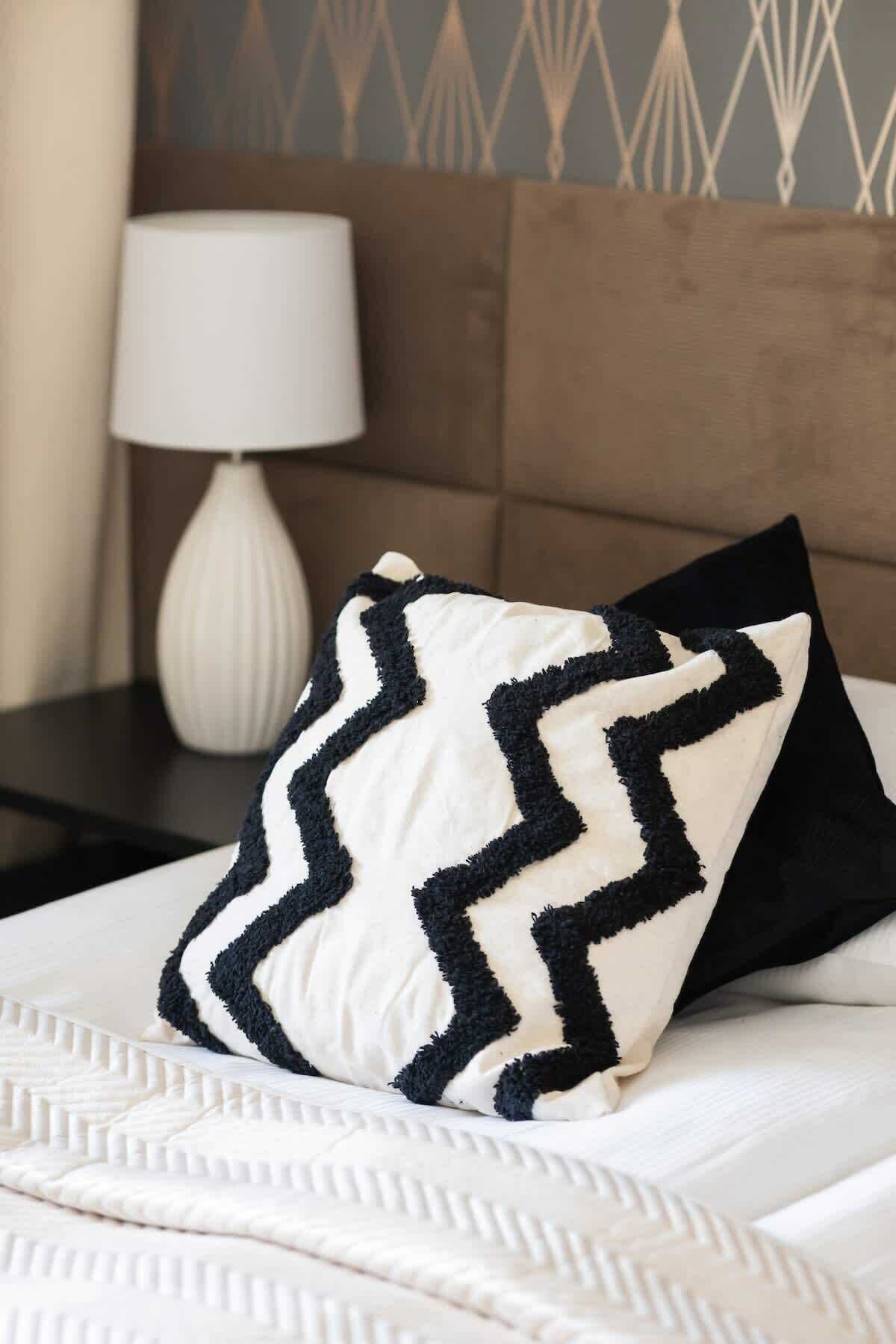 A neatly made bed with a white and black zigzag patterned pillow, a white lamp, and a brown headboard.