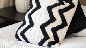 Una cama bien hecha con un almohadón de estampado blanco y negro en zigzag, una lámpara blanca y un cabecero marrón.
