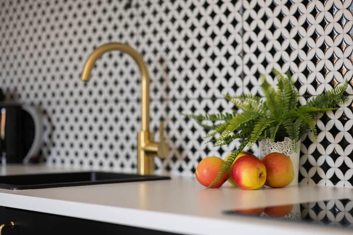 A kitchen counter with a gold faucet, a black kettle, and a vase with green foliage and apples.