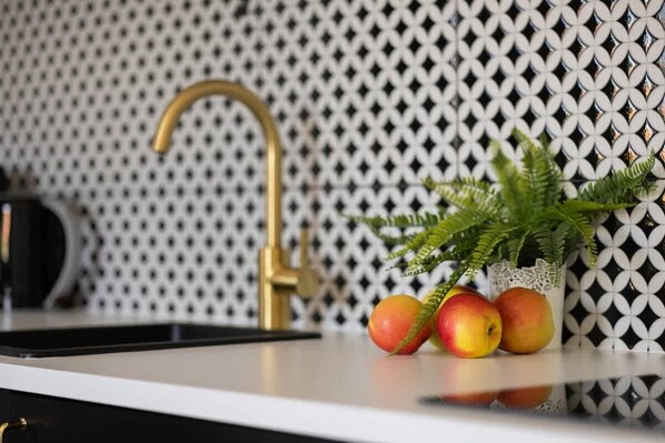 A kitchen counter with a gold faucet, a black kettle, and a vase with green foliage and apples.