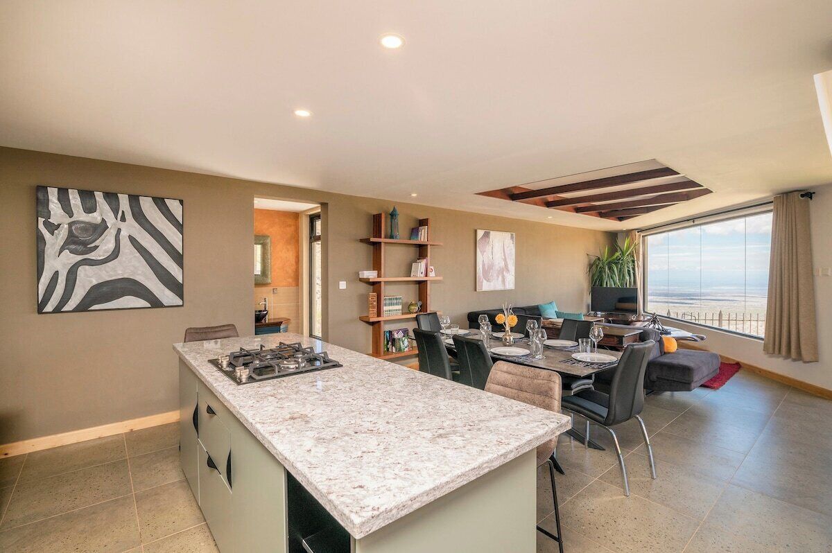 A modern kitchen with a granite countertop, dining area with a table and chairs, a bookshelf, and a large window with a view.