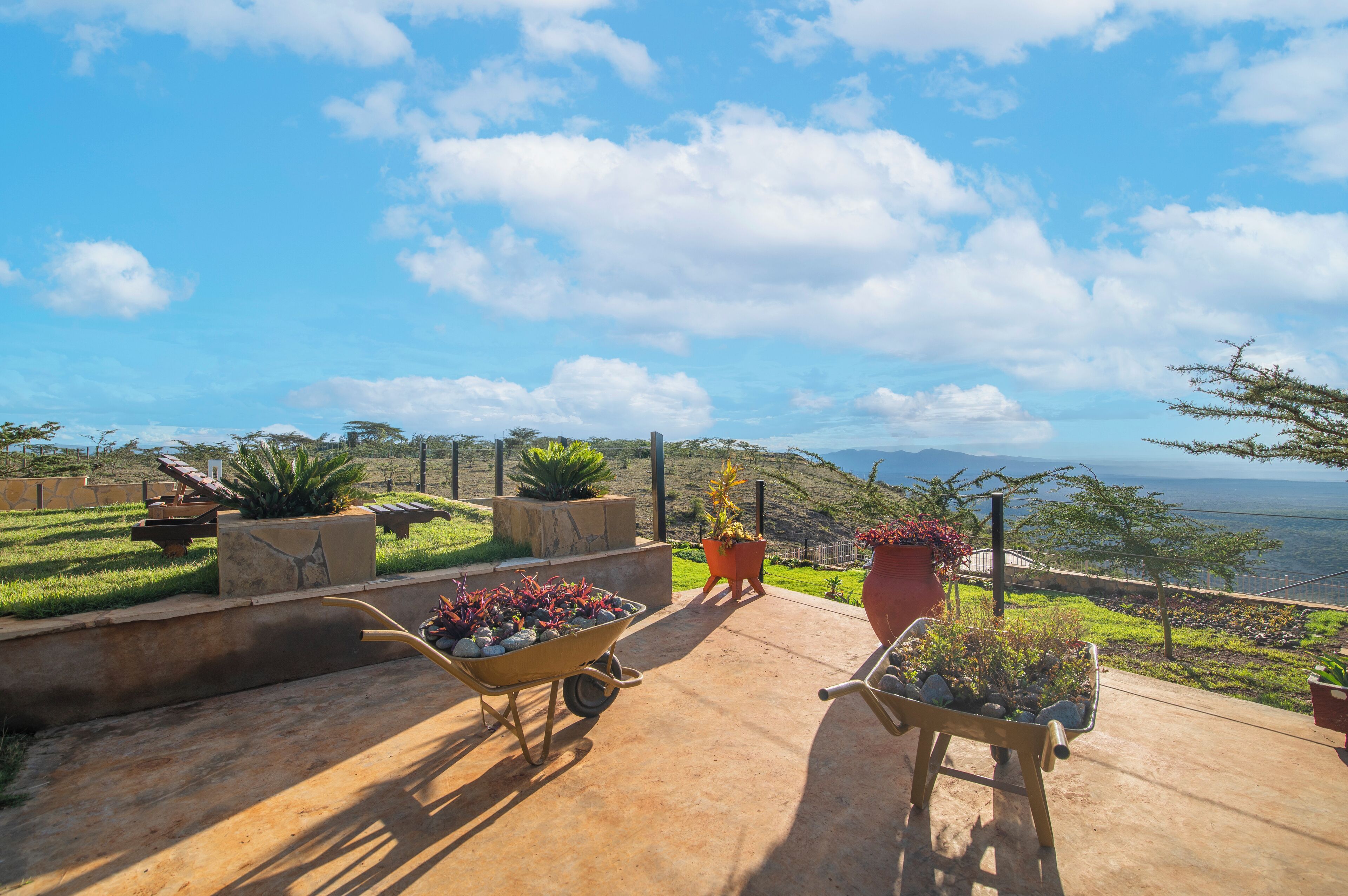 A patio with potted plants and a wheelbarrow, overlooking a scenic view of the ocean and hills.