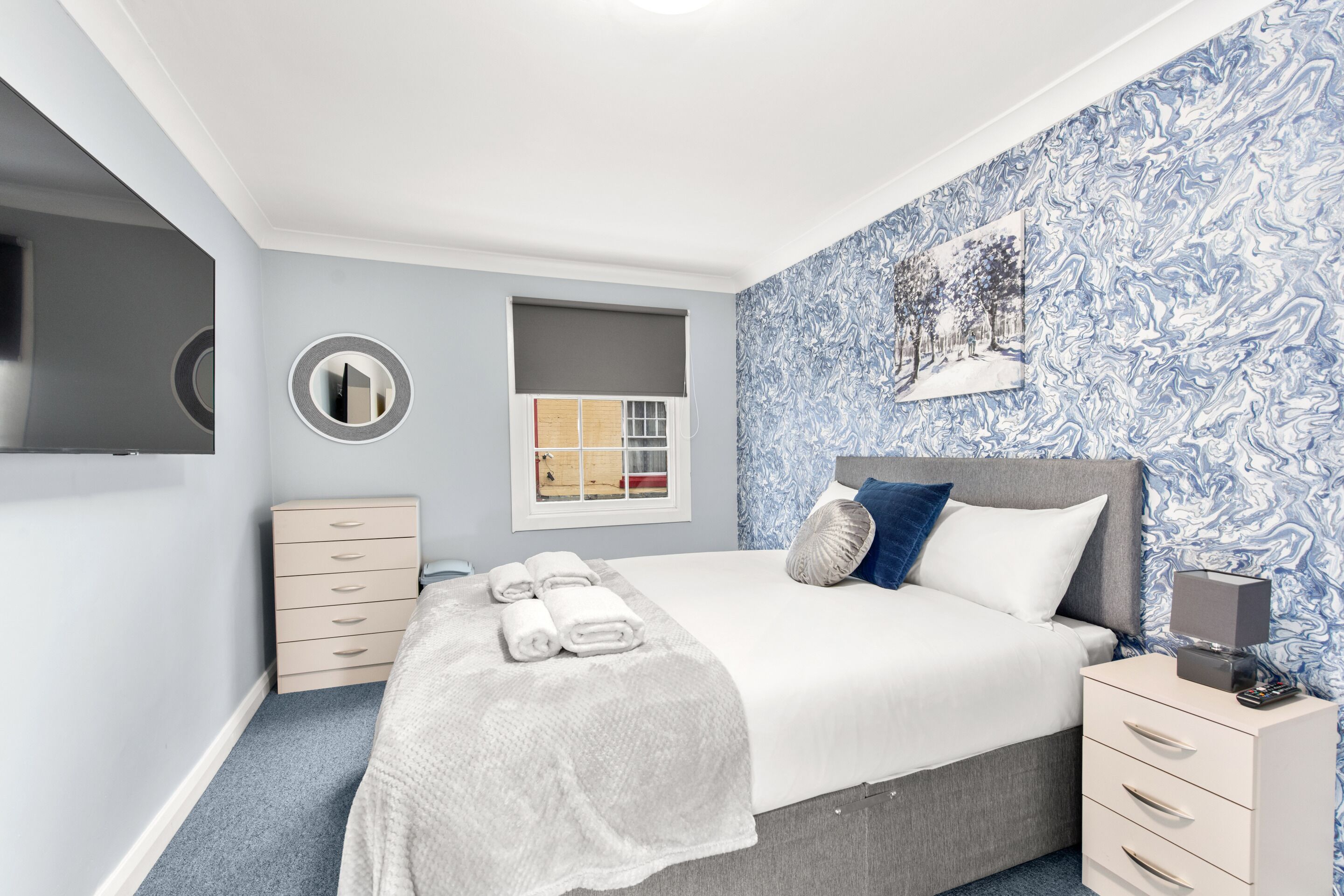 A bedroom with a bed, a bedside table, a dresser, a mirror, a window, and a wall with a blue abstract pattern.
