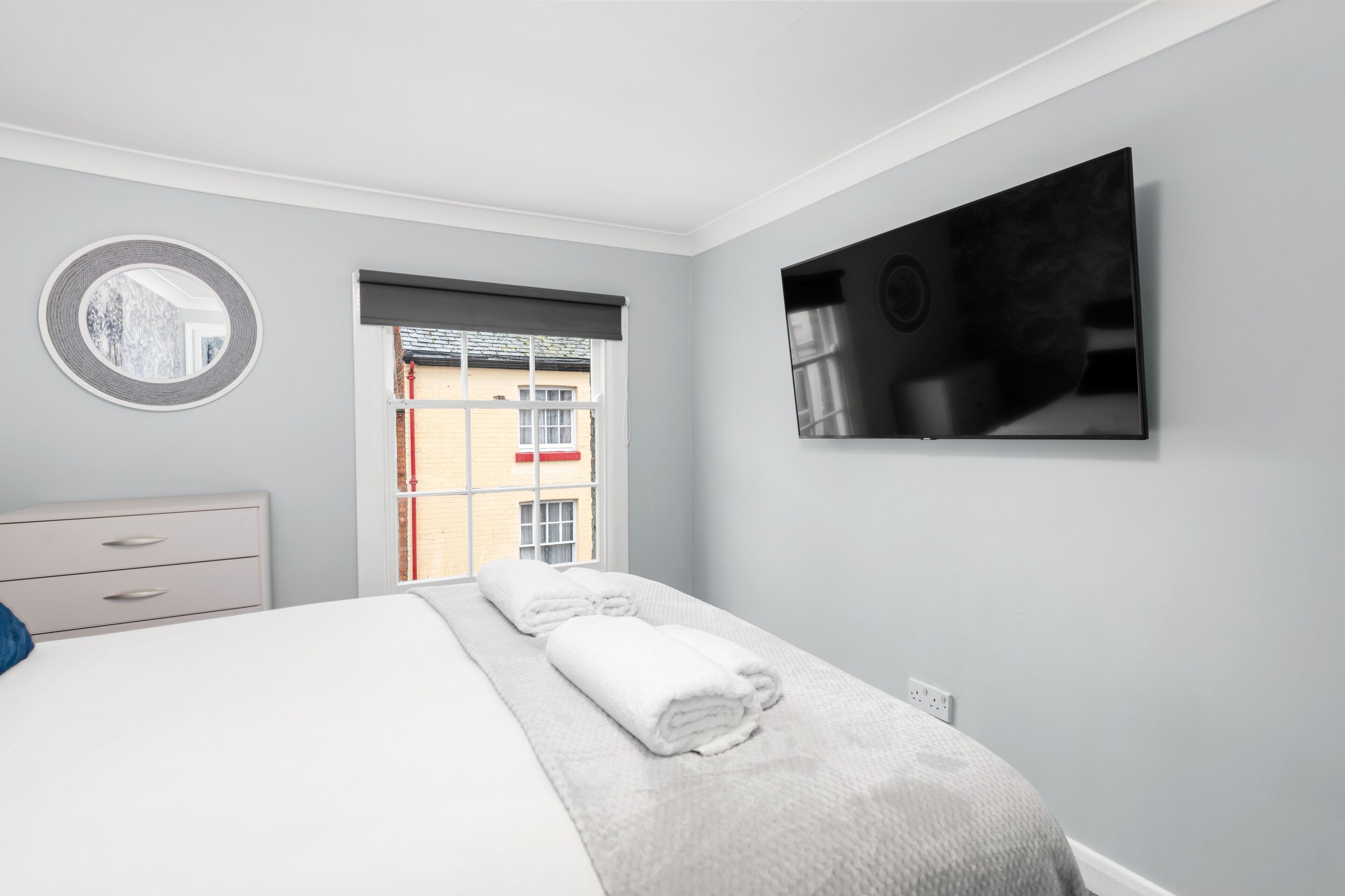 A modern bedroom with a bed, a bedside table, a mirror, a window with a view of buildings, and a flat-screen TV mounted on the wall.