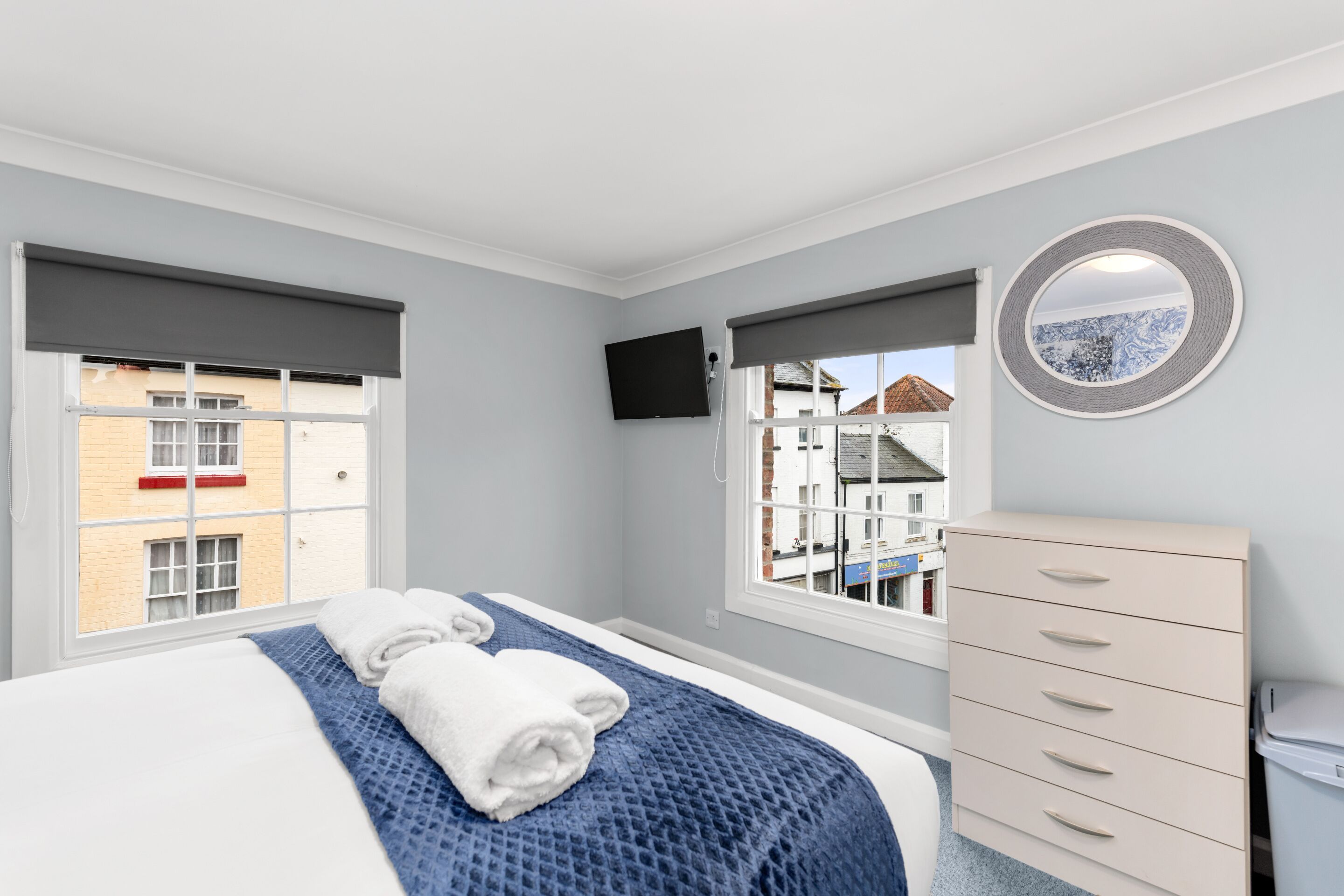 A bedroom with a bed, two windows, a TV, a dresser, and a view of buildings outside.