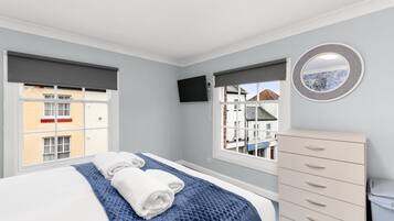 A bedroom with a bed, two windows, a TV, a dresser, and a view of buildings outside.